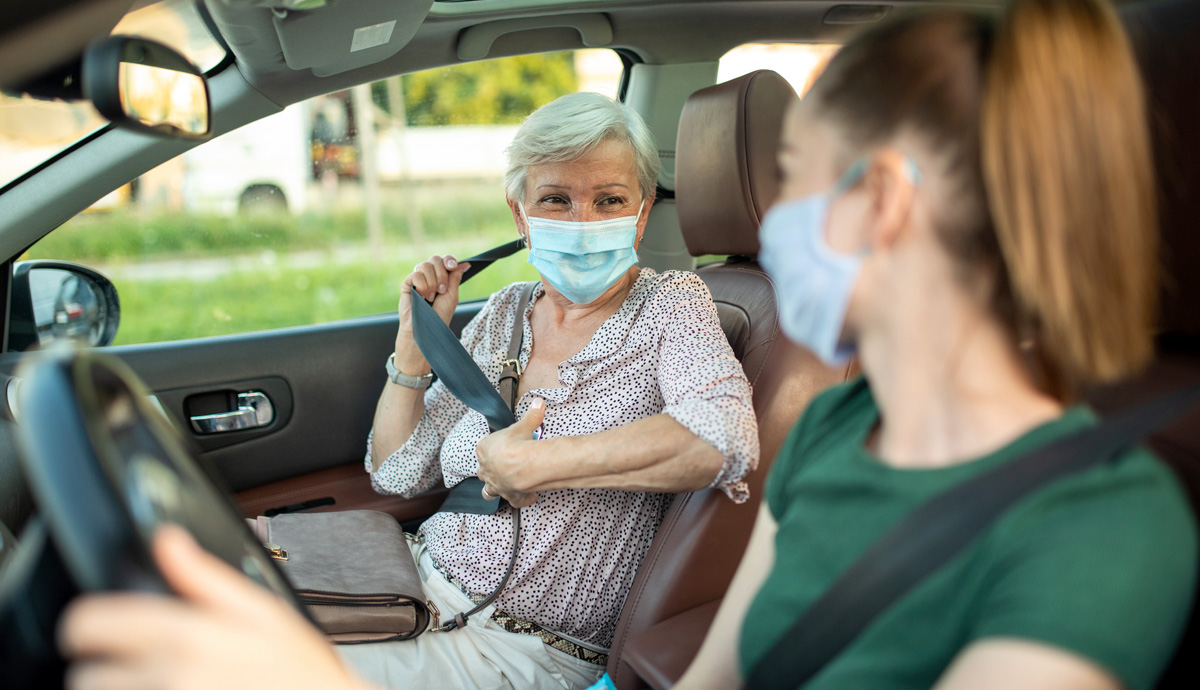 An older woman wearing a mask buckles her seat belt in the passenger seat of a car while looking at a young woman in the driver's seat.