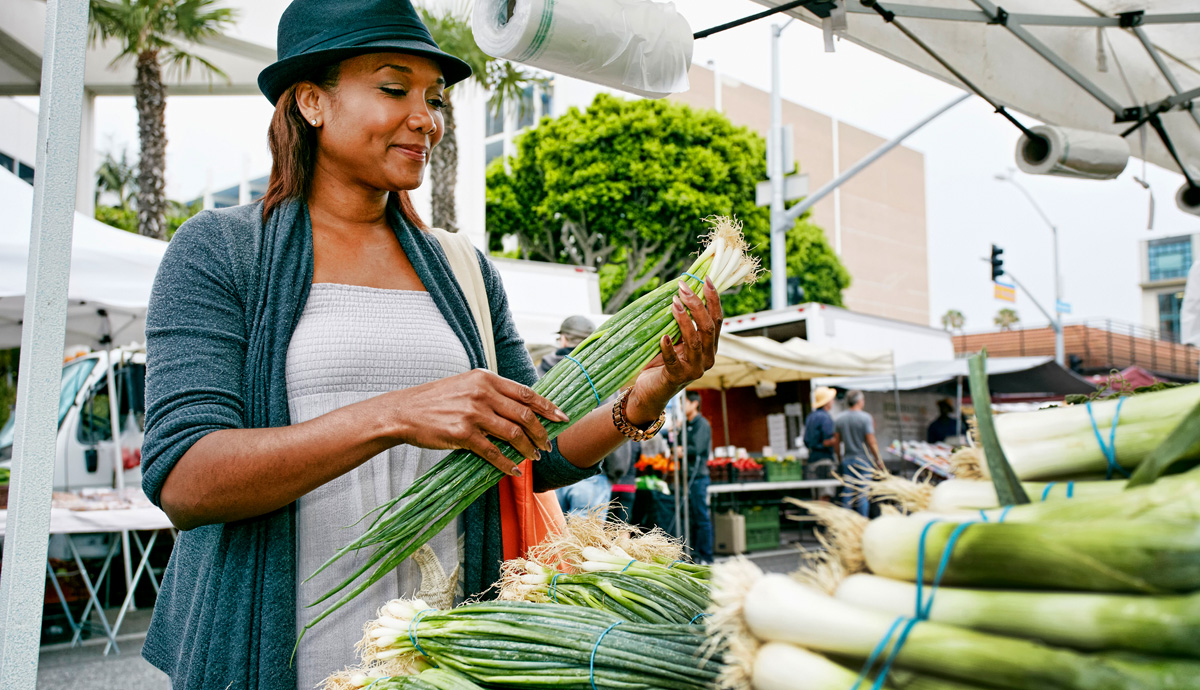 A stylish woman wearing a hat choosing fresh scallions at a farmer's market.