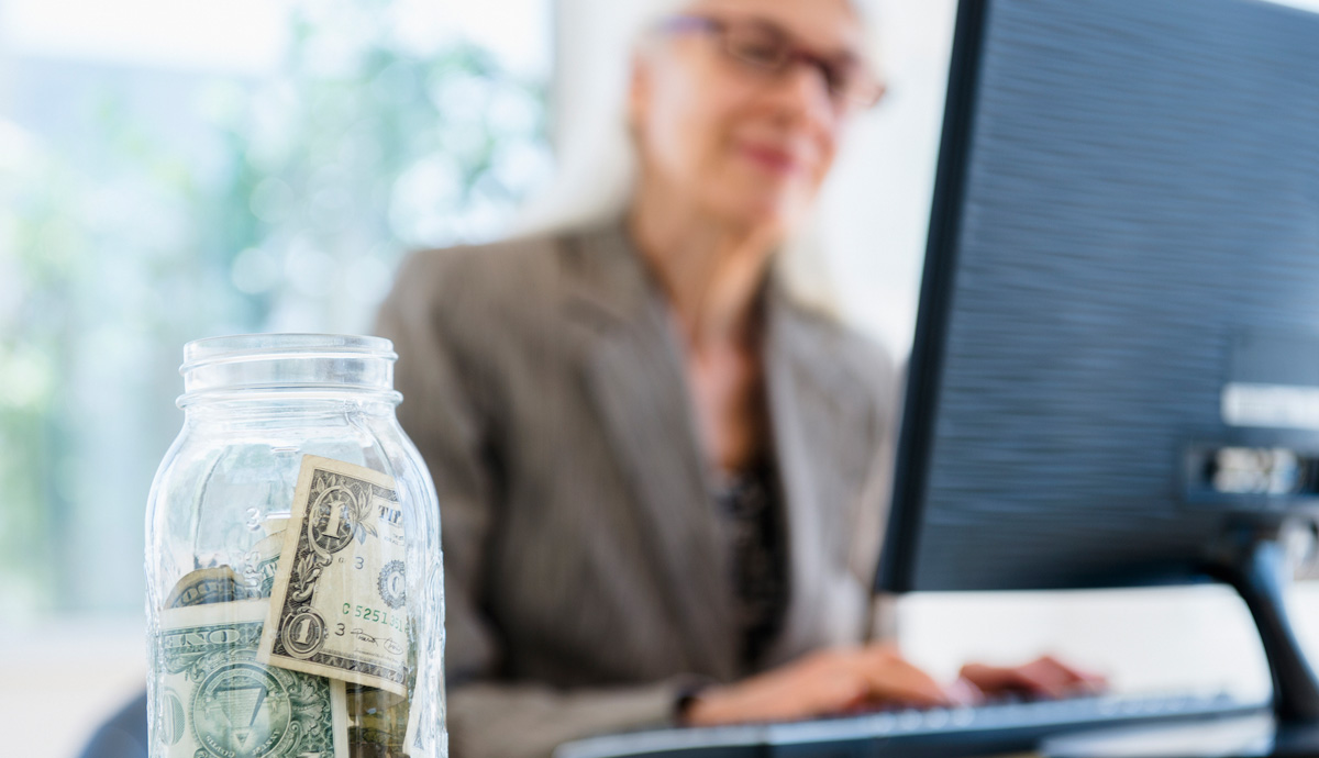 Glass jar filled with money beside a woman on computer in background