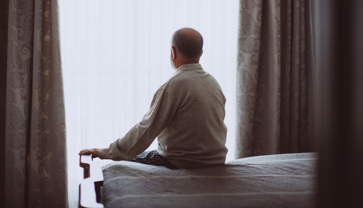 A man sitting on the edge of his bed while looking out a window