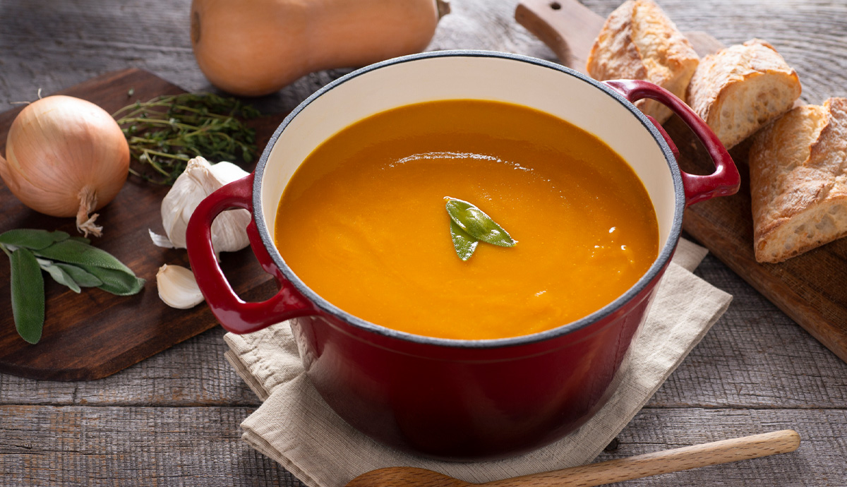 A large colorful pot of pumpkin soup surrounded by ingredients and a sliced baguette