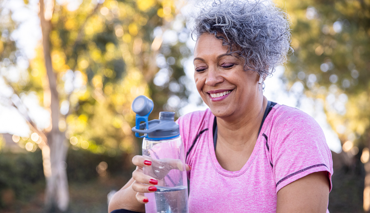 A smiling woman looking at her water bottle after a walk outside.