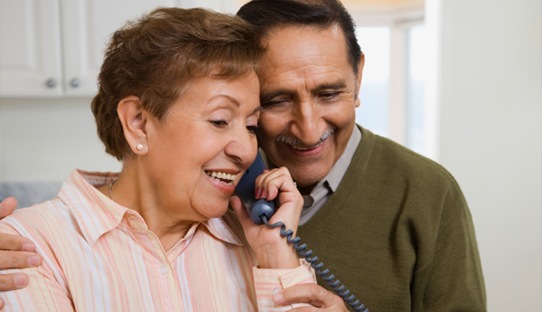 Older Latino couple standing in the kitchen smiling with their heads pressed together listening to the phone.