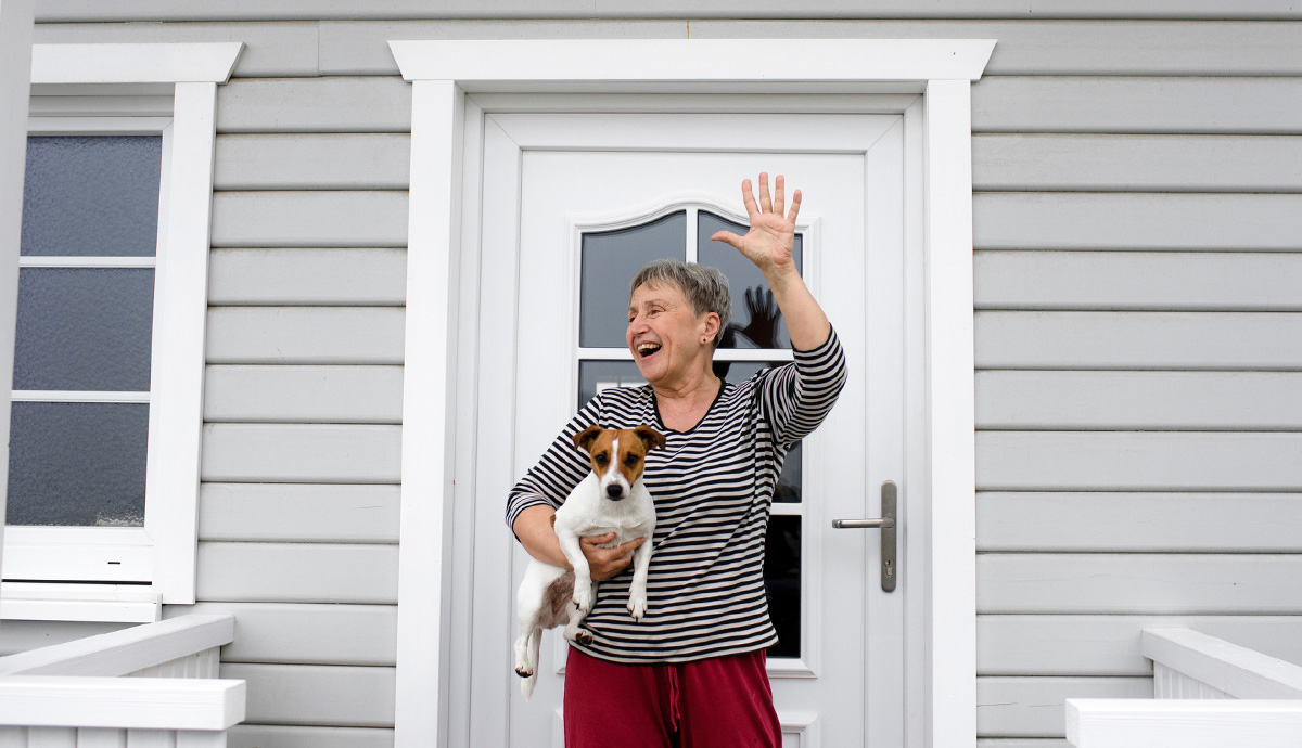 An older woman on her front stoop holding her dog and waving