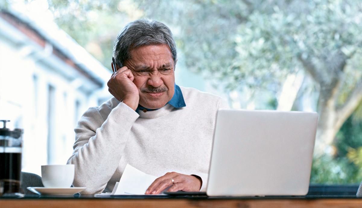 A man wearing a sweater frowns at a laptop while using his arm to prop up his head.
