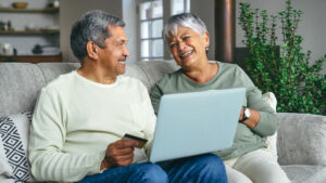 A man and woman sit on a couch together looking at a laptop and smiling.