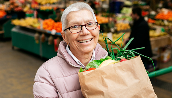 Woman wearing glasses and pink coat, smiling while holding bag of groceries