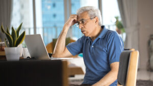 Older adult male sitting at desk with one hand on forehead demonstrating a pensive expression while looking at laptop screen