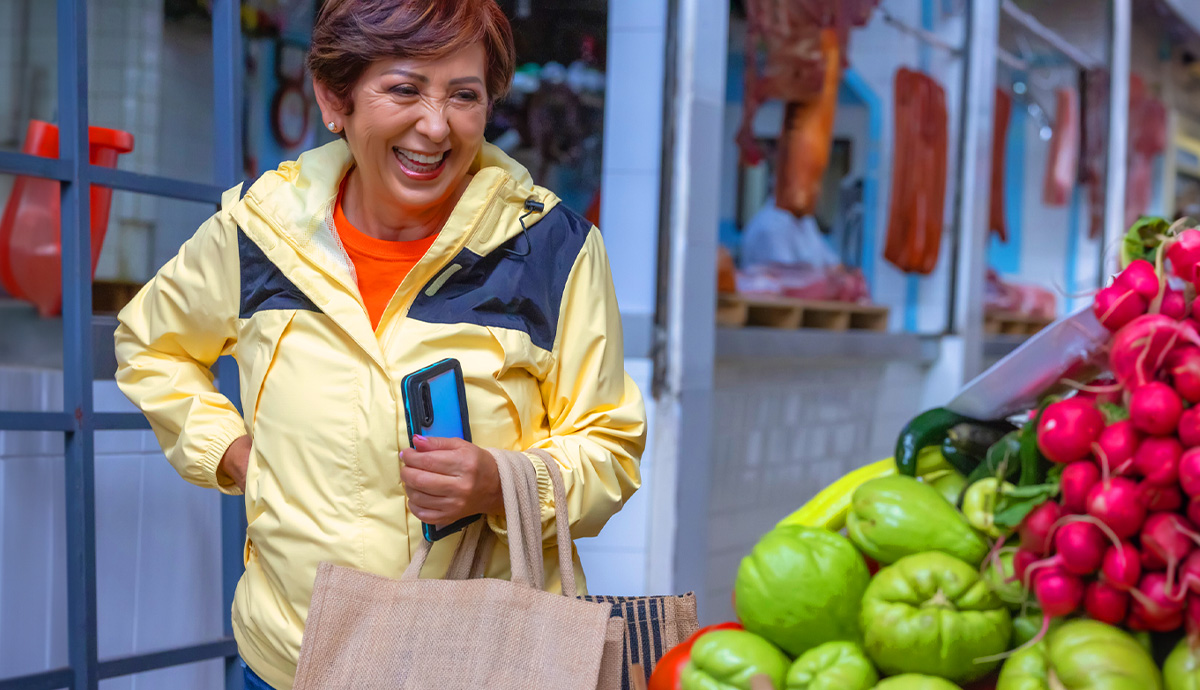 A smiling woman stands in front of a brightly colored vegetable stand in a yellow jacket with brown paper bags on one arm.