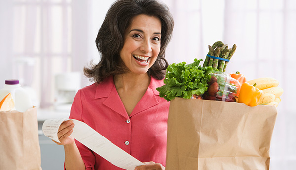 Smiling woman holding receipt while standing in front of bag of groceries inside home.
