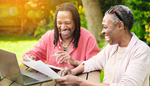 Cheery older man and cheery older woman sitting outside at table with laptop while looking over a document