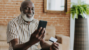 Smiling man sitting wearing striped shirt while holding cell phone and debit card in hands