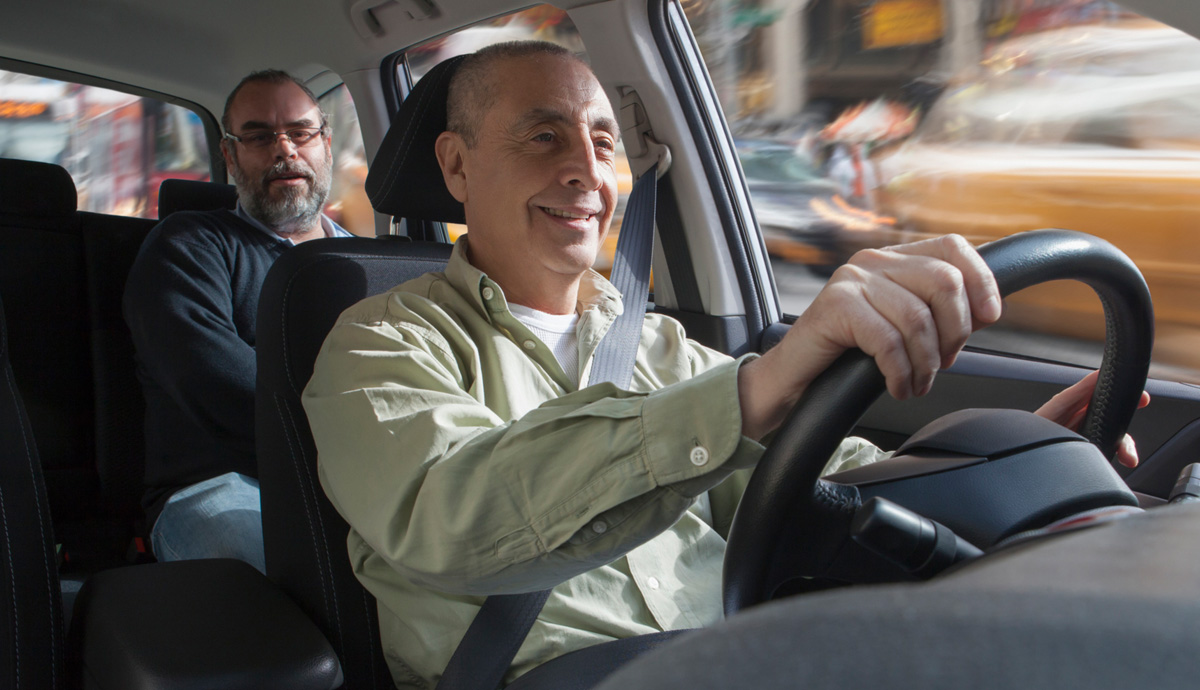 Two men in a car. One is in the driver’s seat with both hands on the steering wheel, the other is riding behind him in the back seat.