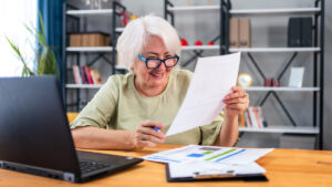 A mature woman sitting at a desk in her home paying bills on her laptop.