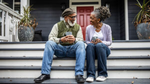 A man and woman sit on their front porch steps gazing at each other while each holds a mug of coffee in their hands.