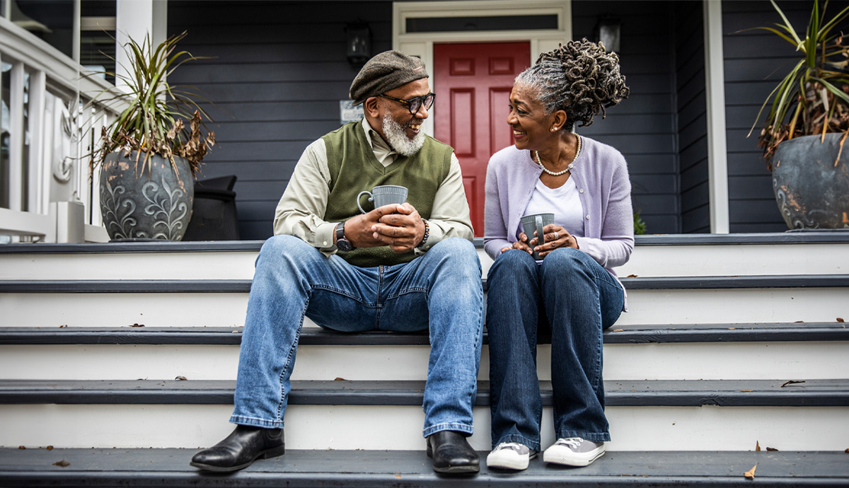 A man and woman sit on their front porch steps gazing at each other while each holds a mug of coffee in their hands.