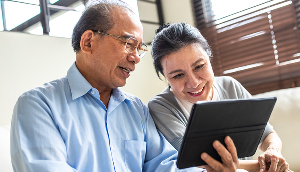 An older couple smiling and sitting on a couch while looking at a smart tablet.