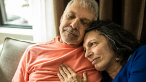 A wife caregiving for her husband lays her head on his shoulder.