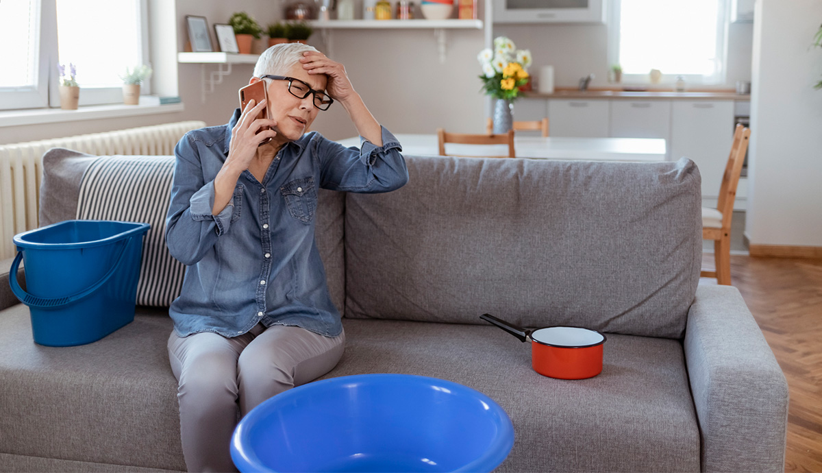 A woman sitting on her couch surrounded by buckets placed to catch leaking water. She is on the phone, looking worried, with her other hand on her head in frustration.