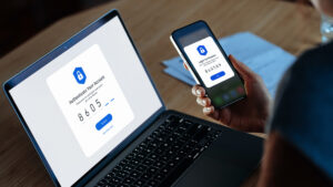 Over the shoulder view of a woman using mobile device with Two-Factor Authentication while logging in securely to her laptop. Over the shoulder view of a woman using a mobile device with Two-Factor Authentication while logging in securely to her laptop.