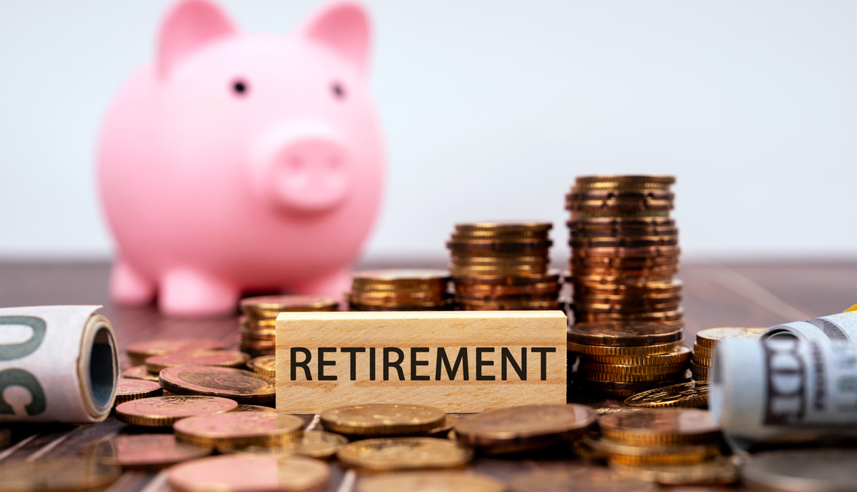 A small wooden block that says Retirement is surrounded by coins and rolled dollar bills with a pink piggy bank in the background.