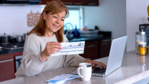 A smiling woman standing at her kitchen island, using a laptop to pay bills.