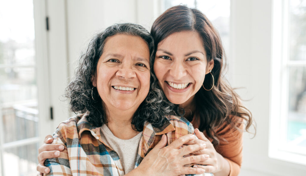 A senior hispanic woman smiles with her adult daughter embracing her.