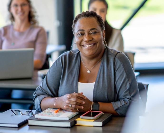 Smiling woman seated at a table with a notebook and smartphone, in a classroom setting.