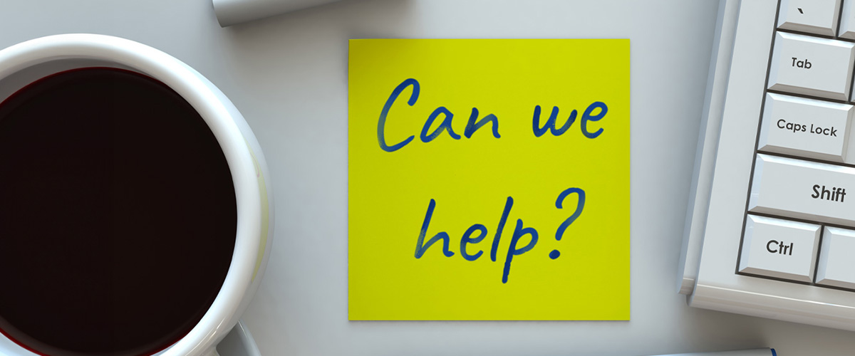 A close-up of a yellow sticky note on a desk that says “Can we help?”