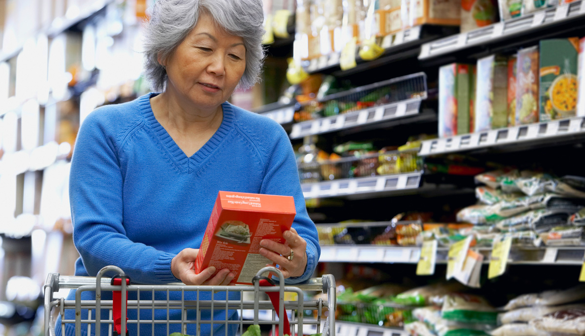 Image of an older woman, standing in a grocery store aisle, looking at an item she pulled off the shelf.