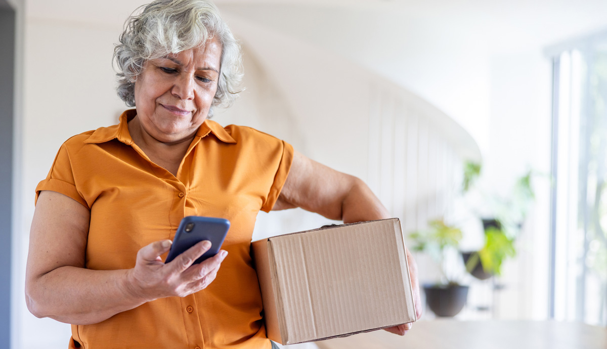 Image of a mature woman in an orange shirt, looking at her phone, holding a moving box