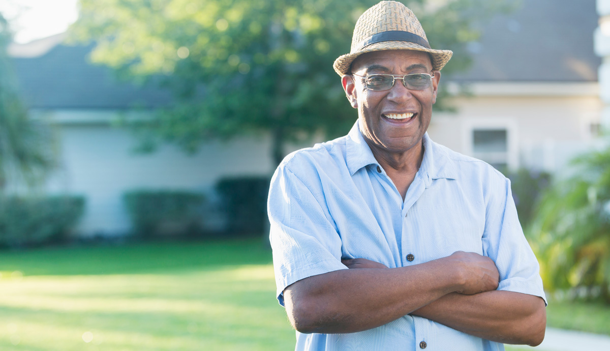 Smiling man wearing hat and eyeglasses with folded arm standing in front yard of home