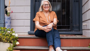 A smiling older woman in an orange top is sitting contentedly on her home's front steps.