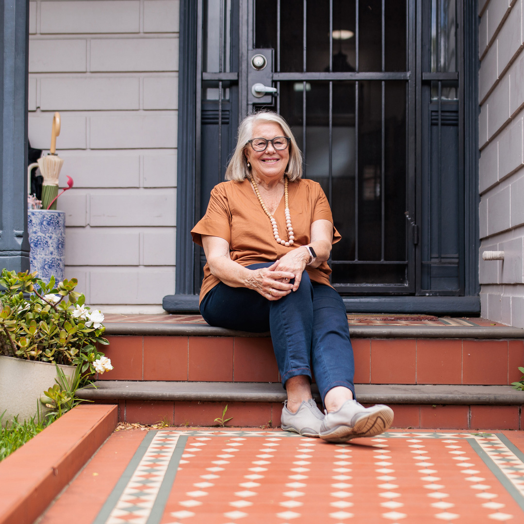 A smiling older woman in an orange top is sitting contentedly on her home's front steps.