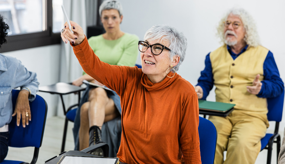 Image of a mature woman, wearing orange, raising her hand in a classroom.