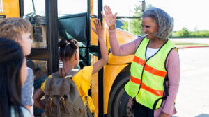 Person in safety vest high fiving children in front of a school bus.